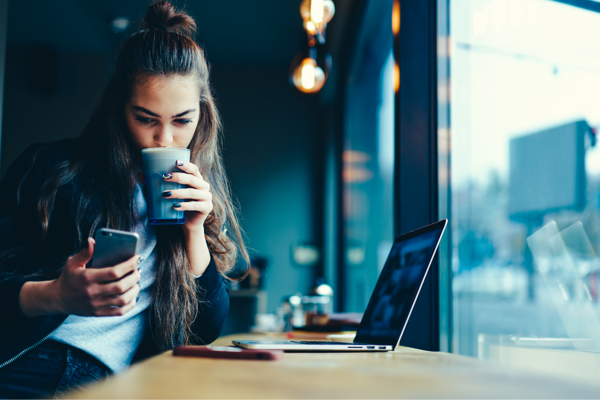 Nuori valkoinen ruskeahiuksinen nainen istuu kahvilassa ikkunapaikalla ja kahvia hörpätessään katsoo älypuhelimen ruutua.